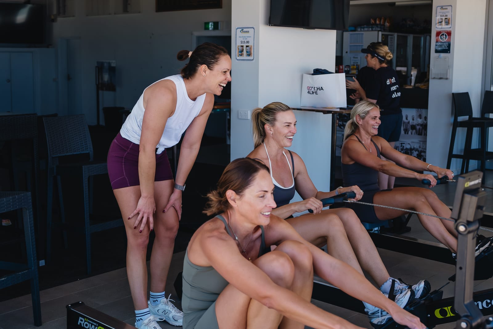 Three women rowing side by side on RowErgs in a club room while a fourth leans in, laughing.