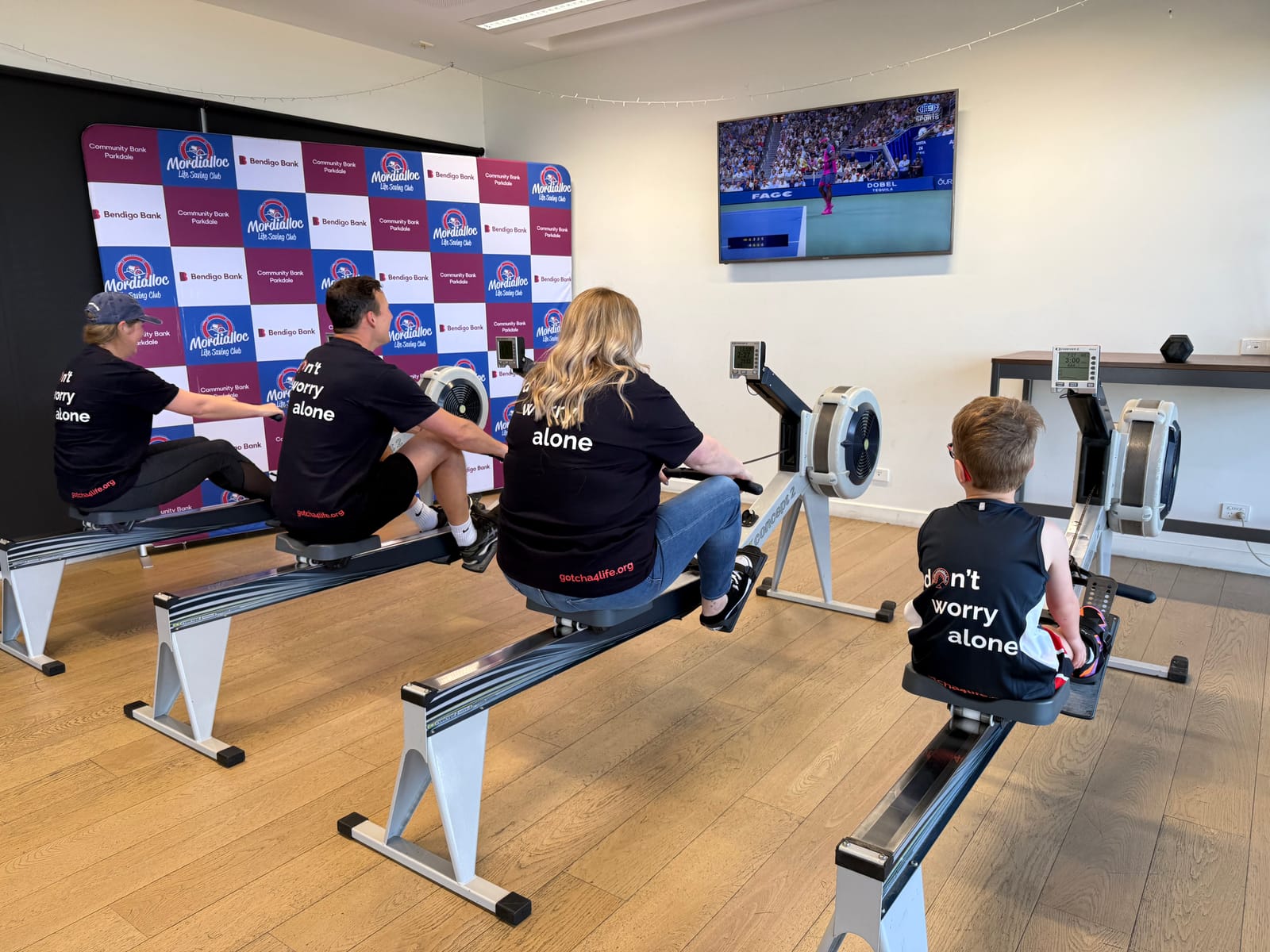 Four people rowing side by side seen from behind, wearing black T-shirts that read don’t worry alone, in front of a Gotcha4Life backdrop.