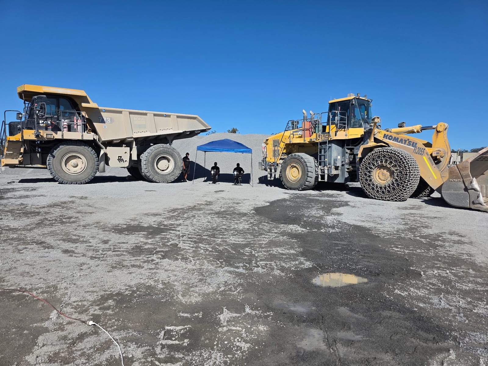 A small blue marquee set up on a mine site between a haul truck and a wheel loader under a clear sky.